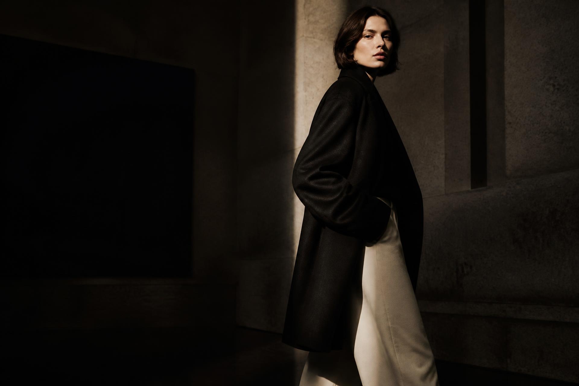Model in a tailored black overcoat and cream trousers in a dim gallery